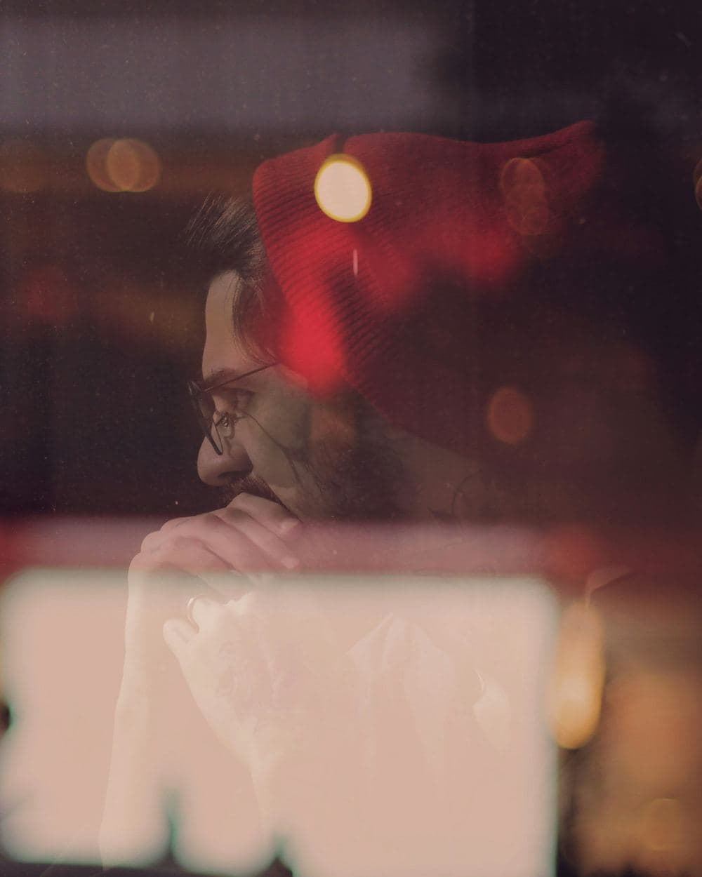 A man seen through a café window at dusk, his presence visible but softened by layered reflections on the glass